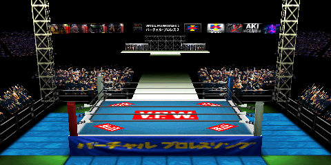 Screenshot of Dome Road, focused on the
ring area. A few items in the far distance: box arts of four of AKI's US-released
N64 wrestling games on the left side; a display in the center featuring a clock
and some text, as well as the AKI Corporation logo; some further logos on the right
hand side. A stage can be seen in the upper center. This leads to the ring area,
though an unlit area separates the two. Some fans are ringside, with light trusses
near the corners. The ring mat design has five 'V.P.W.' logos, each consisting of
a red rectangle and white text. Most of the non-logo portion is teal, but there is
a white border section at the edges. The ring skirt is a darker blue, with yellow
Japanese text. Like Budokan, the outside corner areas without mats are green.
The outside mats are light blue, similar to the ones in AKI Hall.