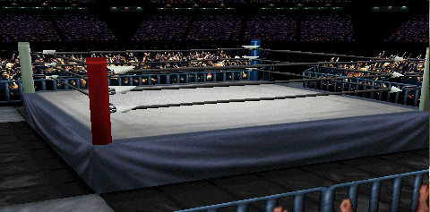 Screenshot of 'USA Dome', focused on
the ring area. Many fans appear in the distance, representing the far seats.
There is also a contingent of fans closer to the ring. Blue guardrails separate
the ringside fans from the action. The ring itself appears on an angle. The primary
mat design is a gray square, bordered by a darker gray edge. This dark gray continues
to the ring skirt design. Outside the ring are four sets of black mats, with the
corners being a lighter blue-ish concrete.